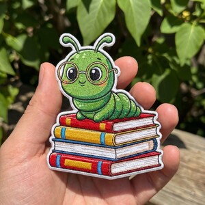 May include: An embroidered patch featuring a bright green cartoon caterpillar with large eyes and round glasses resting on a stack of four colorful books. The books are red, yellow, white, and red with blue accents, all outlined in white thread.