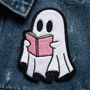 May include: Embroidered patch featuring a white ghost reading a pink book. The ghost has black eyes and a black outline. The patch is on a blue denim jacket.