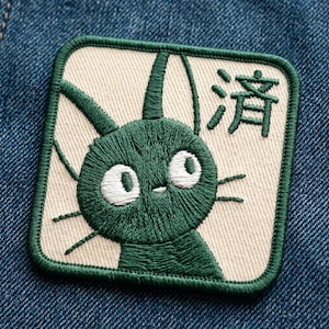 May include: Embroidered patch featuring a green cat-like creature with large white eyes and whiskers. The patch is square with a cream-colored background and a green border. Japanese characters are printed on the top right corner. The patch is on a blue denim background.