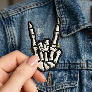 May include: A black and white embroidered patch in the shape of a skeleton hand making the rock and roll hand gesture. The patch has a black border and is pinned to a denim jacket.