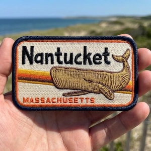 May include: A rectangular embroidered patch with a dark blue border, featuring a cream-colored background, a brown sperm whale illustration, and horizontal orange and brown stripes. The text reads Nantucket in black and MASSACHUSETTS in orange.