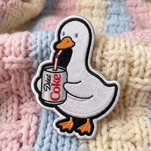 May include: An embroidered patch featuring a white duck with an orange beak and feet. The duck is holding a silver soda can with the text Diet Coke printed in red and black, with a pink straw extending from the can to the duck's beak.