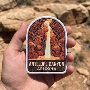 May include: An embroidered patch with a white border featuring a depiction of Antelope Canyon in shades of brown, orange, and tan. A vertical beam of light descends through the canyon walls. The text ANTELOPE CANYON ARIZONA is in white.