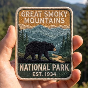 May include: A rectangular embroidered patch featuring a black bear walking through a forest landscape with blue mountains in the background. The text reads GREAT SMOKY MOUNTAINS NATIONAL PARK EST. 1934 in white lettering against tan and green backgrounds.