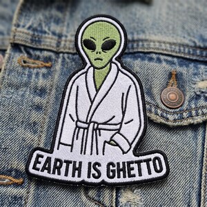 May include: Embroidered patch featuring a green alien wearing a white robe. The alien has black eyes and a serious expression. The text "EARTH IS GHETTO" is displayed in white letters on a black background.