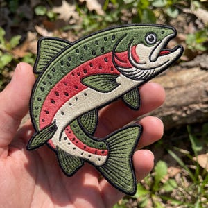 May include: An embroidered patch shaped like a rainbow trout fish. The fish features a dark green back with black spots, a vibrant red stripe along its side, and a cream-colored belly. The design is outlined in black thread with detailed fins.