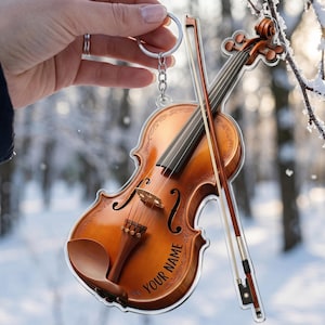 May include: A violin-shaped keychain with a bow, featuring a light brown finish and the text "YOUR NAME" in a curved font. The keychain is held by a hand against a blurred winter forest backdrop.