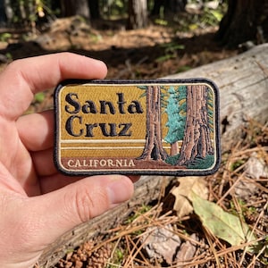 May include: A rectangular embroidered patch featuring a mustard yellow background with the black text Santa Cruz and the word CALIFORNIA in white. The design includes brown redwood trees and a green pine tree with a black stitched border.
