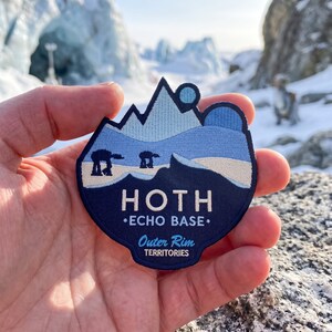 May include: A shield-shaped embroidered patch featuring a snowy mountain landscape in shades of light and dark blue. The patch displays the text HOTH ECHO BASE and Outer Rim TERRITORIES, along with two dark silhouettes of four-legged walking vehicles.