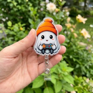 May include: An acrylic badge reel featuring a white ghost character wearing an orange beanie and holding a black video game controller with colorful buttons. The ghost has large black eyes and a small smiling mouth, presented as a flat graphic.