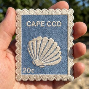 May include: A rectangular embroidered patch shaped like a postage stamp with a scalloped edge. The patch features a light blue background with the text "CAPE COD" at the top, a beige seashell graphic in the center, and "20c" printed at the bottom.