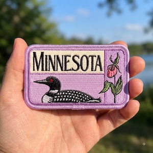 May include: A rectangular embroidered patch with a light purple border. The patch features the text MINNESOTA in black letters on a cream background, a black and white loon with a red eye, and a pink lady slipper flower on a green stem.