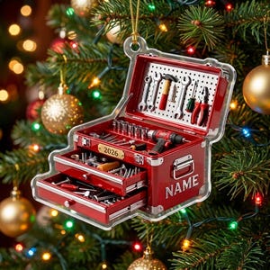 May include: A red, tiered toolbox ornament featuring a pegboard lid with miniature tools, two open drawers filled with hardware, a gold plaque labeled 2026, and the word NAME printed on the side, all encased in a clear acrylic frame.