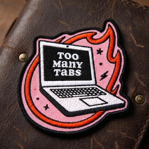 May include: Embroidered patch featuring a laptop with the text "Too Many Tabs" on the screen. The laptop is surrounded by a pink circle and orange flames, with small star and lightning bolt accents. The patch is on a brown leather surface.