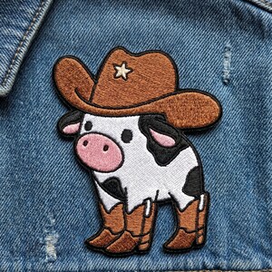 May include: Embroidered patch featuring a cartoon cow wearing a brown cowboy hat with a star and brown cowboy boots. The cow has a white body with black spots, a pink nose, and black eyes. The patch is on a blue denim background.