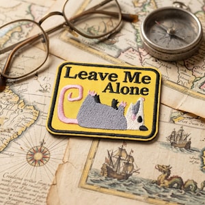 May include: A yellow patch with the text "Leave Me Alone" and an embroidered opossum. The opossum is gray, white, and black, with a pink tail. The patch is on a vintage map, with glasses and a compass.