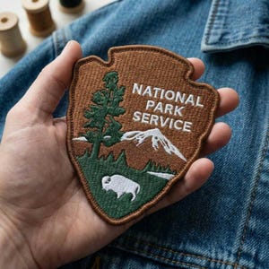 May include: A brown and green National Park Service patch in the shape of an arrowhead. The patch features a tree, mountains, and a bison. The words "NATIONAL PARK SERVICE" are embroidered in white.