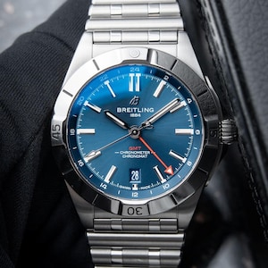 May include: A stainless steel Breitling Chronomat watch with a blue dial. The watch features a date display at the 6 o'clock position and a GMT function. The watch has a silver bezel and a stainless steel bracelet.