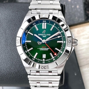 Op de afbeelding: Een Breitling Chronomat horloge van roestvrij staal met een groene wijzerplaat. Het horloge heeft een datumweergave, een GMT-functie en een zilveren armband. De wijzerplaat heeft lichtgevende wijzers en markeringen. Het horloge is een luxe uurwerk.