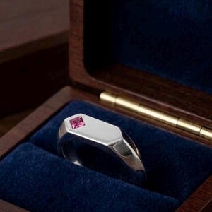 May include: A silver signet ring featuring a single, small, square-cut pink gemstone set into the flat, polished top surface. The ring has a smooth, metallic finish and a solid band, presented within a dark wooden box with a blue velvet interior.