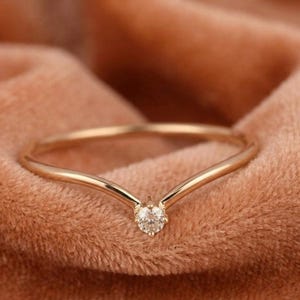 May include: A gold-colored ring with a V-shaped design. The ring features a small, round, clear gemstone at the center of the V. The ring is set against a soft, peach-colored background.