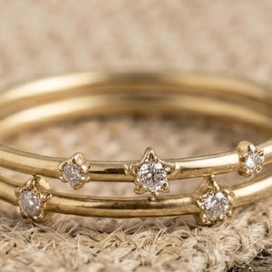 May include: Three gold-colored rings stacked together, each adorned with small, sparkling star-shaped diamonds. The rings are set against a neutral, textured background, highlighting the jewelry's delicate design.