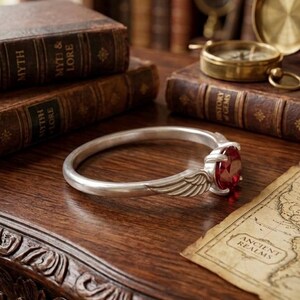 Op de afbeelding: Een zilveren armband met een rode edelsteen en vleugelaccenten. De armband ligt op een houten oppervlak met antieke boeken, een kompas en een kaart met het opschrift "Ancient Realms". Het ontwerp van de armband is gedetailleerd en elegant.