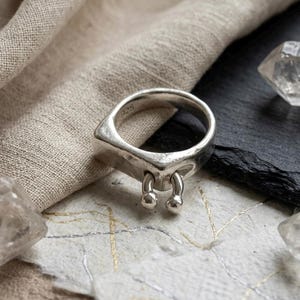 May include: A silver ring with a square top and two dangling ball charms. The ring is made of a shiny metal and is set against a neutral background.