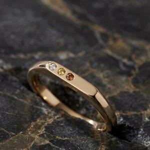 May include: A gold band ring featuring three small, round, flush-set gemstones arranged in a row. The stones are graduated in colour, transitioning from a clear white diamond-like stone to a yellow stone, and finally to a warm amber-brown stone.