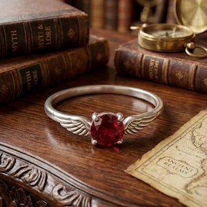 Op de afbeelding: Een zilveren ring met een grote, ronde, rode edelsteen en vleugelvormige accenten. De ring ligt op een houten oppervlak met antieke boeken, een kompas en een kaart op de achtergrond. Het ontwerp van de ring roept een fantasiethema op.