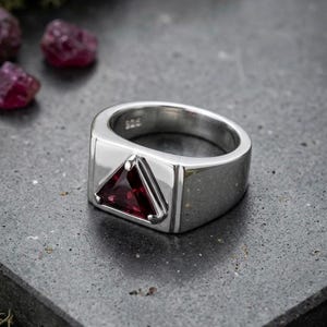 Pode incluir: Um anel de prata com uma pedra preciosa vermelho-escura de corte triangular no centro de uma banda polida. O interior da banda está claramente marcado com o selo 925, indicando a composição metálica desta peça de joalharia.