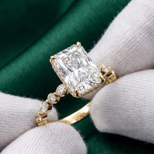 May include: A gold ring featuring a large rectangular radiant-cut centre stone held by four prongs. The band is adorned with a series of smaller round-cut stones set along the shank, with additional tiny stones lining the basket setting.