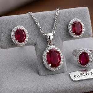 May include: A silver jewellery set comprising a necklace, earrings, and a ring. Each piece features a large oval ruby gemstone encircled by small white stones. The set is displayed on a grey velvet surface. The text "925 Silver & Ruby Set" is visible.
