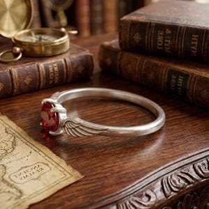 Op de afbeelding: Een zilveren armband met een grote, ovale, rode edelsteen. De armband heeft een vleugelontwerp. De sieraden worden getoond op een donkere houten tafel met antieke boeken, een kaart en een kompas.
