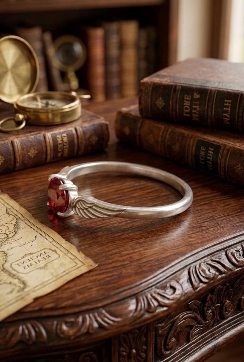 Op de afbeelding: Een zilveren armband met een grote, ovale, rode edelsteen. De armband heeft een vleugelontwerp. De sieraden worden getoond op een donkere houten tafel met antieke boeken, een kaart en een kompas.