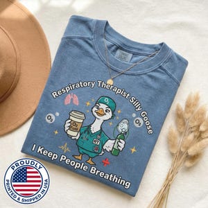 May include: A blue Comfort Colors sweatshirt with a graphic of a goose in a medical uniform holding a coffee cup and oxygen tank. Text reads "Respiratory Therapist Silly Goose" and "I Keep People Breathing". A gold necklace and a "Proudly Printed & Shipped in USA" sticker are also visible.