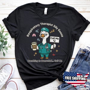 May include: Black t-shirt with a cartoon goose dressed as a respiratory therapist. The shirt features the text "Respiratory Therapist Silly Goose" and "Breathing Is Overrated... Call Me". The goose holds a coffee cup and a sign that says "Call Me".