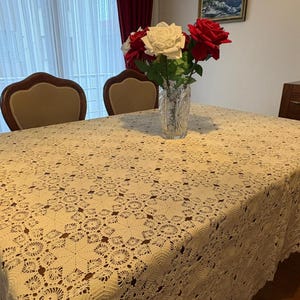 May include: A cream-colored, crocheted tablecloth covers a wooden table. A clear glass vase holds a bouquet of red and white roses. Two chairs with brown upholstered seats are visible. A framed landscape painting hangs on the wall.