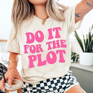 May include: Cream-colored t-shirt with the phrase "DO IT FOR THE PLOT" in bright pink, bold, retro-style lettering. The shirt is paired with black and white checkered shorts.