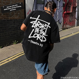 May include: A black short-sleeve t-shirt featuring white graphic text on the back that reads Trust in the Lord and Proverbs 3:5 in a stylized font. The shirt is paired with blue denim shorts. A black sign above displays Best for: DTG, DTF, Sublimation, Screen Printing, and more.