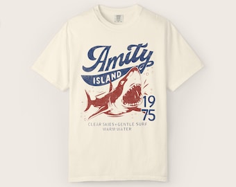 May include: Cream-colored t-shirt featuring a distressed graphic of a shark with the text "Amity Island 1975". The design includes the phrases "Clear Skies + Gentle Surf Warm Water" in a vintage style.