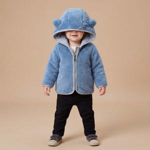 May include: A light blue fleece jacket with a hood featuring bear ears. The jacket has a zipper closure and a gray trim. The child is wearing black pants and blue sneakers.