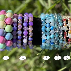 May include: Five beaded bracelets in various colors. The bracelets include pink, purple, blue, and white beads. Some bracelets have star-shaped beads. The bracelets are displayed on a black surface.