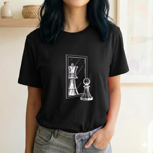 Könnte beinhalten: Ein schwarzes Kurzarm-T-Shirt mit einer weißen Strichgrafik auf der Brust. Das Design zeigt einen Schachbauern vor einem rechteckigen Spiegel, wobei die Spiegelung eine Schachkönigin darstellt.