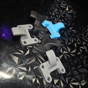 May include: A selection of 3D-printed brackets in grey, black, and blue. Each bracket features a distinctive design with mounting holes and a hook-shaped projection. The brackets are arranged on a textured, dark surface.