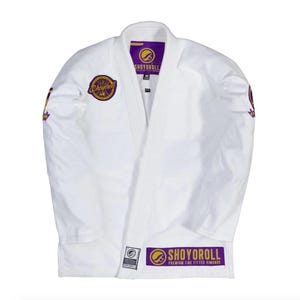 Puede incluir: Uniforme blanco de artes marciales con detalles morados y dorados. El uniforme presenta la marca "SHOYOROLL" en el pecho, las mangas y el dobladillo inferior. Fabricado con un material resistente, está diseñado para entrenamiento y competición.