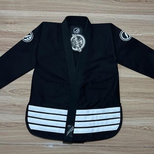 May include: Black martial arts uniform with white accents. The uniform features a black jacket with white stripes at the bottom and white circular patches on the sleeves. A tag with text is visible on the jacket.
