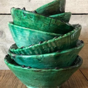 May include: A stack of six handmade, green ceramic bowls. The bowls have a textured, glazed finish and a slightly irregular shape. The rims are decorated with small, dark accents. The bowls are stacked on a wooden surface.