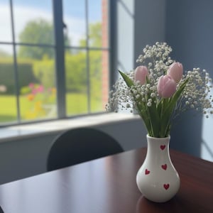 May include: A white vase with red heart designs holds a bouquet of pink tulips and white baby's breath. The vase sits on a dark wood table near a window with a view of a green yard.