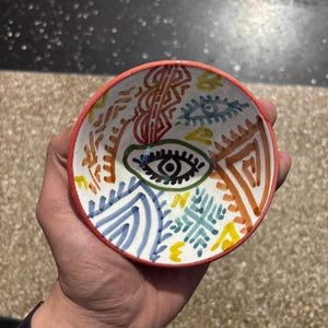 Puede incluir: Un cuenco pequeño y redondo de cerámica con borde rojo e interior blanco. El cuenco está decorado con diseños abstractos coloridos, incluyendo ojos, líneas y formas geométricas en azul, naranja, amarillo y verde. El cuenco está en una mano.